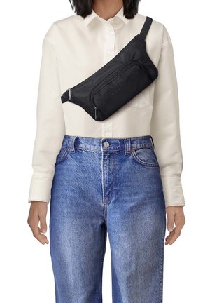 Czarna torba na ramię z tkaniny o fakturowanej powierzchni i zamykanymi na zamek kieszeniami, noszona na ciele przez białą koszulę i niebieskie dżinsy.