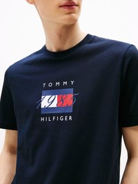 T-shirt in cotone di un blu navy con una grafica rettangolare che riporta "TOMMY," "1985," e "HILFIGER" in bianco e rosso. Collo rotondo classico.