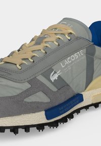 Γκρι και μπεζ αθλητικό παπούτσι Lacoste με κίτρινα κορδόνια, μπλε εσωτερικό, υφασμένο πέλμα και λογότυπο της εταιρείας με κείμενο στο πλάι.