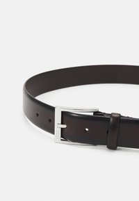 Cinturón de cuero marrón con textura suave, que presenta una hebilla de metal plateado y varios agujeros de ajuste. Grabado visible en la hebilla.