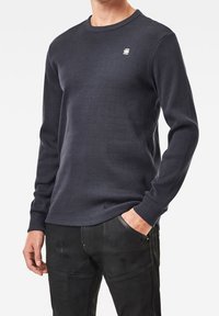 Långärmad, ribbad marinblå sweatshirt med rund halsringning och liten logga på bröstet, matchad med svarta byxor med fickor och sömmar.