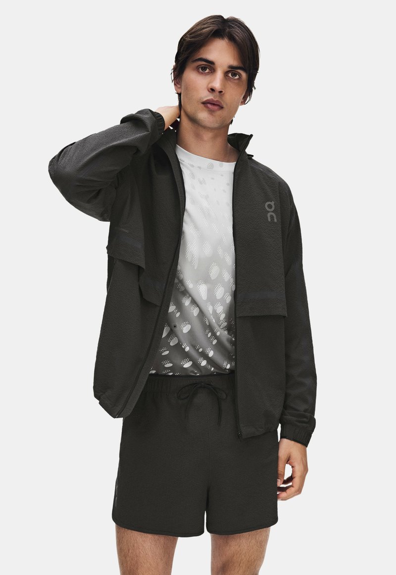 Jeune homme portant une veste noire zippée, une chemise blanche à motifs et un short noir, debout avec une main derrière le cou.