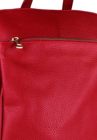Sac à dos en cuir rouge avec une surface texturée, doté d'une poche zippée dorée à l'avant et de coutures contrastées. Pas de quincaillerie visible.