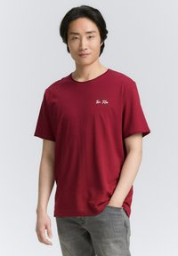 Rød bomulds t-shirt med rund hals, korte ærmer og et lille hvidt logo på venstre bryst, parret med lyse grå jeans.