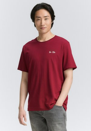 T-shirt en coton rouge avec un col rond, des manches courtes, et un petit logo blanc sur la poitrine gauche, associé à un jean gris clair.