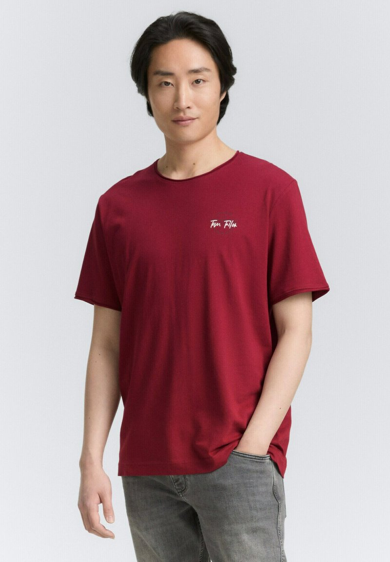 Rød bomulds t-shirt med rund hals, korte ærmer og et lille hvidt logo på venstre bryst, parret med lyse grå jeans.