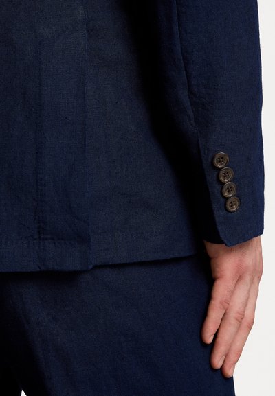 Navy linen bleiseri, jossa on tyylikäs istuvuus, teksturoitu pinta ja neljä ruskeaa nappia hihassa. Läheltä kuvattu näkymä hihasta ja olkapäästä.