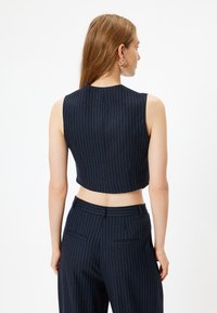 Marineblaues Cropped Top mit vertikalen weißen Nadelstreifen, hohem Kragen und ärmellosem Design, kombiniert mit passenden Nadelstreifen-Hosen. Glatte Textur.