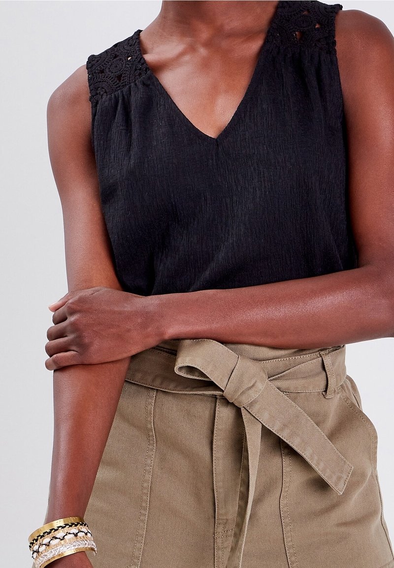 Haut noir sans manches avec col en V et détails en dentelle sur l'épaule. Porté avec un pantalon taille haute beige clair ceinturé à nouer.