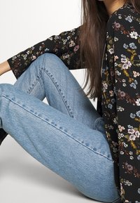 Kvinna sitter med benen i kors iförd ljusblå jeans och en svart blommig långärmad tröja mot en vit bakgrund.