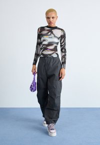 Transparent long-sleeve top with abstract print, paired with black baggy pants. She holds a purple patterned bag. Sneakers feature pink accents.