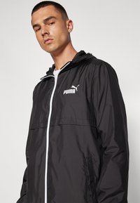 Chaqueta ligera negra con capucha, que presenta una cremallera blanca y un logo de Puma blanco en el lado izquierdo del pecho. Material suave y resistente al agua.
