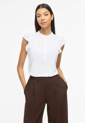 Žena s dlhými tmavými vlasmi, ktorá nosí bielu blúzku s krátkym rukávom a hnedé nohavice s vysokým pásom, stojí s rukami v vreckách na bielom pozadí.