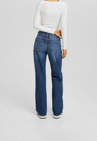 Wide-leg blue denim jeans with a high waist, visible stitching, and two back pockets. Paired with a fitted white long-sleeve top.