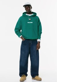 Ung mand iført en grøn hættetrøje med teksten "STWD STUDIO", brede blå jeans, brune støvler, sølvhalskæde, hvid kasket og kædearmbånd.