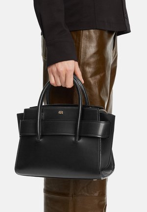 Persona sosteniendo un bolso de cuero negro con asas dobles, vistiendo pantalones de cuero marrón y una camiseta negra de manga larga.