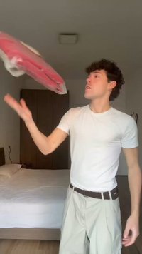 Junger Mann in weißem T-Shirt und beigen Hosen wirft einen roten, in Plastik eingewickelten Gegenstand in einem Schlafzimmer mit Bett und Kleiderschrank.