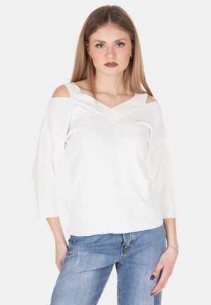 Giovane donna con lunghi capelli biondi, che indossa un top bianco con le spalle scoperte e jeans blu, in piedi davanti a uno sfondo chiaro e uniforme.