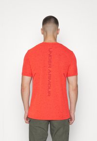 Camiseta atlética corta de manga roja con un patrón de cuadros texturizado; presenta el logotipo "UNDER ARMOUR" impreso verticalmente en la parte posterior.