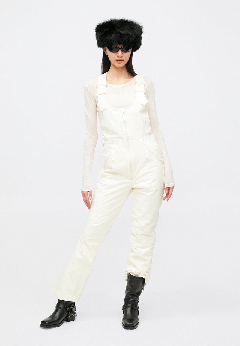 Tuta bianca isolata con bretelle regolabili e tasche con cerniera, abbinata a una maglia trasparente a maniche lunghe e cappello di pelliccia nero. Include stivaletti neri alla caviglia.