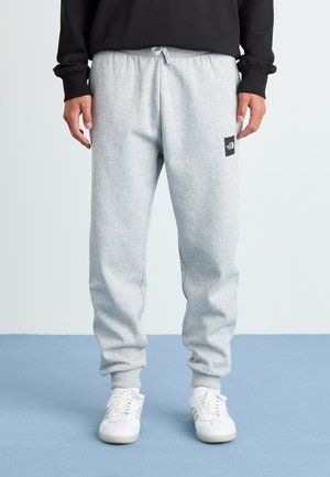 Man wearing light gray sweatpants with The North Face logo on the thigh, white sneakers, and a black long-sleeve top, standing on blue floor.