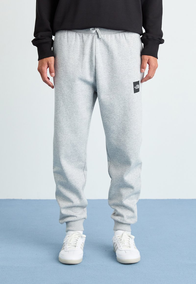 Mand iført lysegrå sweatpants med The North Face-logoen på låret, hvide sneakers og en sort langærmet trøje, står på et blåt gulv.