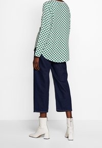 Grünes Polka-Dot-Oberteil mit langen Ärmeln und abgerundetem Saum, kombiniert mit dunkelblauen, weiten, cropped Jeans und weißen, mit Reißverschluss versehenen, Absatzstiefeletten.