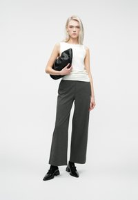 Femme debout dans un large pantalon gris, top sans manches blanc, chaussures noires, tenant une pochette noire en tissu contre un fond blanc uni.