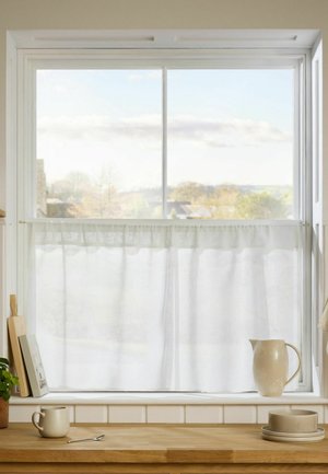 White half-curtain on kitchen window with wooden countertop below holding a mug, spoon, pitcher, plates, and cutting boards.