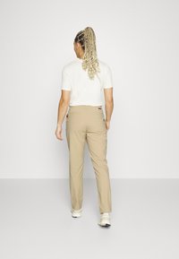 Beige, lätta byxor med rak passform, med bakfickor och en subtil logotypdetalj. Kombinerat med en enkel vit t-shirt.