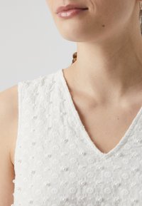 Balta be rankovių viršutinė dalis su V formos iškirpte, tekstūruotu gėlėtu raštu, akcentuota mažomis perlamutrinėmis karoliukais, demonstruojanti švelnų, elegantišką įvaizdį.