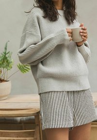 Pull gris clair côtelé avec des manches tombantes, associé à un short rayé noir et blanc ; tenant une tasse beige. Plante en arrière-plan.