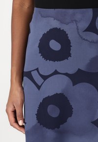 Ženská ruka leží vedle modré sukně s velkými tmavomodrými abstraktními květinovými vzory.