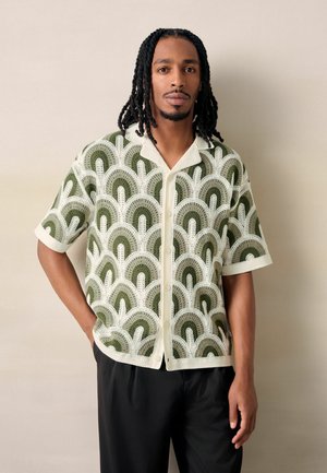 Mężczyzna z dredami, ubrany w krótki sweter szydełkowy z zielonymi i kremowymi wzorami w kształcie łuków oraz czarne spodnie, stojący na jednolitym tle.