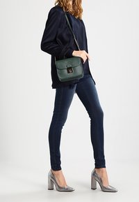 Dark blue long-sleeve top paired with fitted dark jeans. Green structured handbag with a clasp. Silver high-heeled shoes with a block heel.