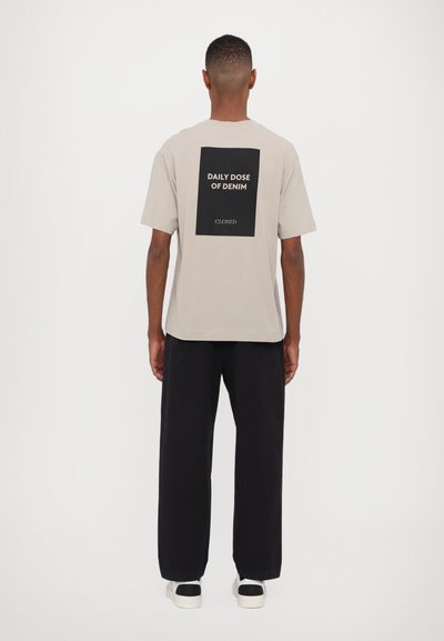 Beige bomulls t-skjorte med et svart grafisk trykk som leser "DAILY DOSE OF DENIM" på baksiden, parret med løse sorte bukser og hvite joggesko.