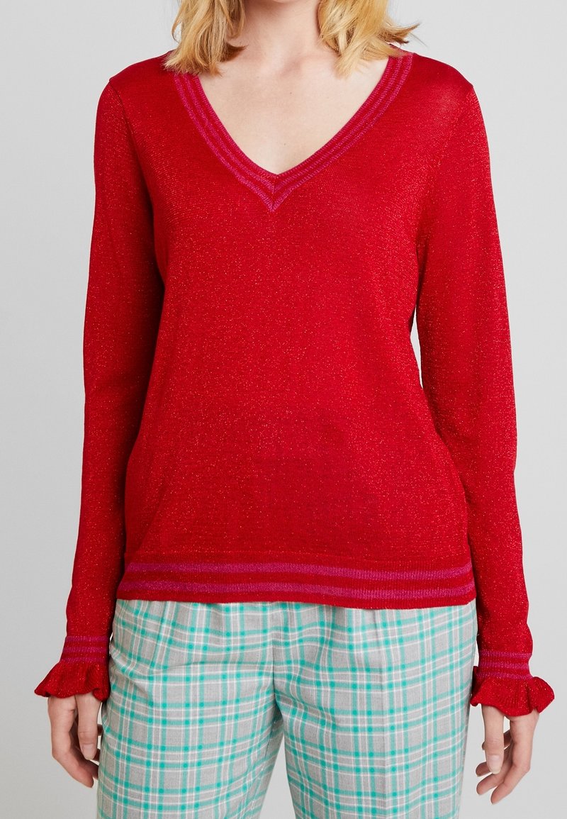 Femme portant un pull à col en V rouge scintillant avec des poignets à volants et un pantalon à carreaux bleu sarcelle et blanc, sur un fond gris uni.