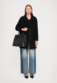 Manteau en laine noir avec de gros boutons, associé à un sac à main noir structuré. Jean en denim large bleu clair avec un dégradé et un ourlet effiloché.