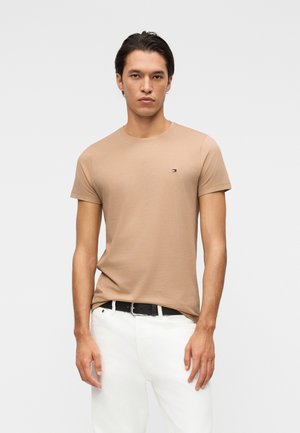 Junger Mann steht, trägt ein beiges kurzärmeliges T-Shirt mit kleinem Logo auf der Brust und weiße Hose mit schwarzem Gürtel, neutraler Hintergrund.