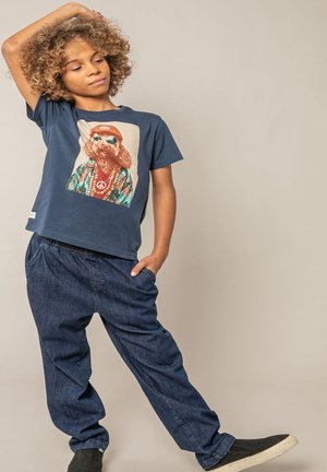 Enfant aux cheveux bouclés portant un t-shirt graphique bleu, un pantalon bleu marine et des chaussures noires à enfiler, posant avec une main sur la tête et l'autre dans une poche.
