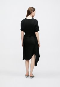 Juoda suknelė su marškinėlių tipo viršumi, draperuota medžiaga liemenį, asimetrinė apačia ir lygi tekstūra. Dėvima su dirželiais papuoštomis sandalais.