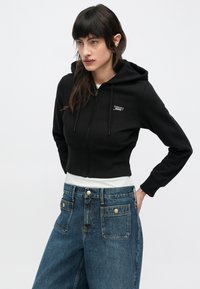 Sudadera negra recortada con cremallera, bolsillo frontal y cordón, con un pequeño logo blanco. Combinada con jeans de mezclilla azul de talle alto.