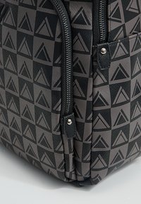 Zaino con motivo geometrico nero e grigio con accenti triangolari, dotato di doppie cerniere e tessuto testurizzato. Forma rettangolare compatta.