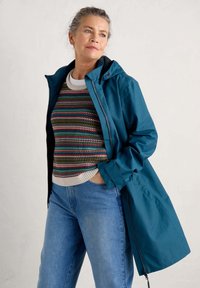 Teal vattentät jacka med huva, dragkedja och sidofickor, lager på lager över en flerfärgad randig tröja och ljusblå jeans.