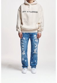 Cremefarbener Hoodie mit dem "2Y STUDIOS"-Logo, kombiniert mit abgenutzten blauen Jeans und weißen Turnschuhen. Der Hoodie verfügt über eine Fronttasche und einen lockeren Schnitt.