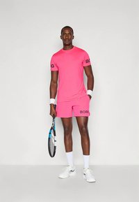 Άνδρας με φωτεινά ροζ αθλητικά ρούχα που κρατάει μια ρακέτα τένις, φορώντας λευκά αθλητικά παπούτσια και βραχιολάκια, στέκεται μπροστά σε απλό φόντο.