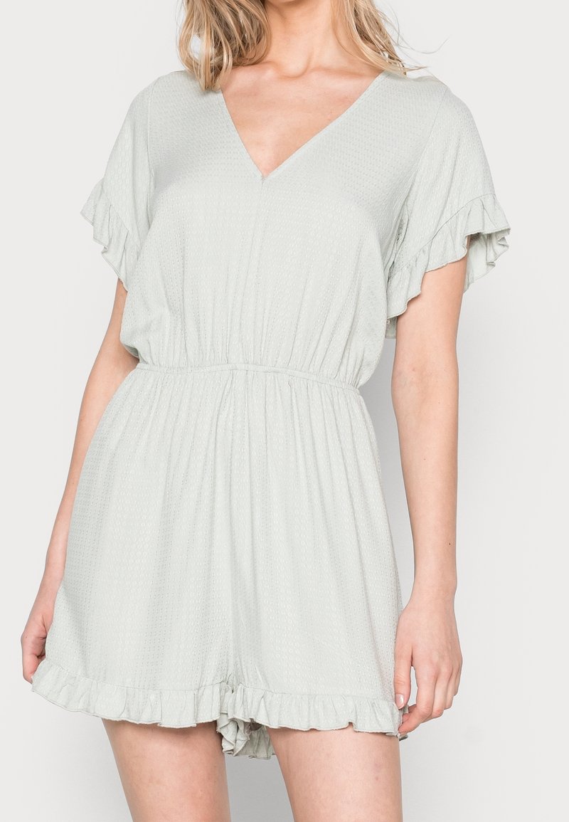 Světlý šedý overal s výstřihem do V, volánkovými rukávy, elastickým pasem a texturovanou látkou. Má vzdušný tvar a volánkový lem.