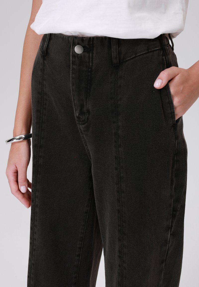Zwarte denim high-waisted broek met een rechte pijp, voorzien van zijzakken en een metallic knopdetail in de taille. Gleffige textuur.
