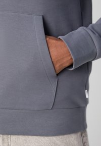 Sivi pulover z sprednjo žepno, rebrastimi manšetami in robom, izdelan iz mehkega bombažnega materiala z gladko teksturo.