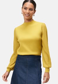Pull jaune avec des manches ballon côtelées, corps ajusté et col montant, associé à une jupe en denim foncé. Texture lisse, sans motifs visibles.